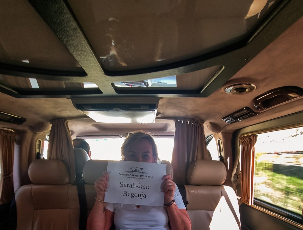SJ sitting in a van in Goreme, Cappadocia, holding up a newspaper with the name "sarah-jane begonja" printed on it, with windows showing a