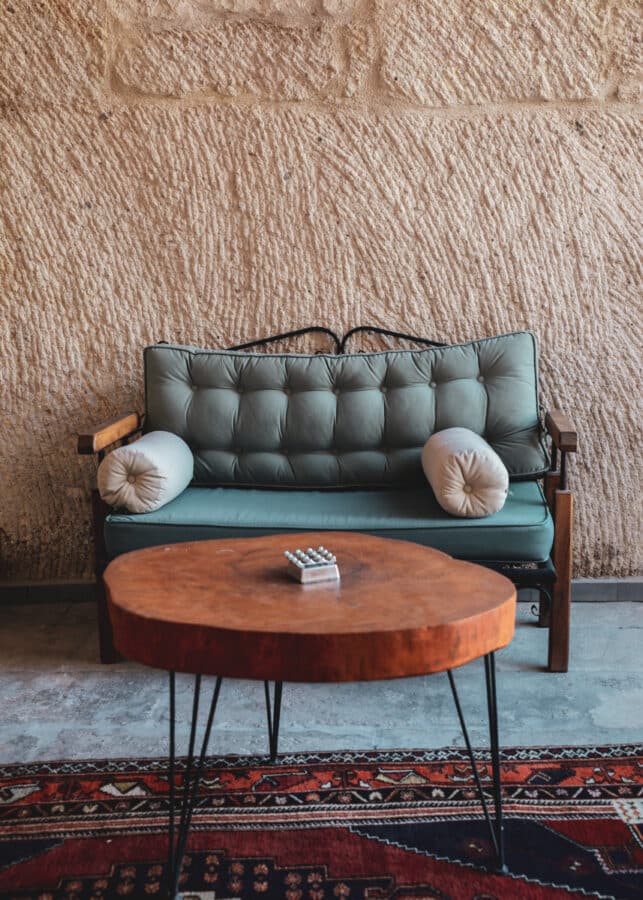 A green tufted sofa with round side cushions and a round wooden coffee table on a colorful rug, against an earthen wall in Goreme, Cappadocia.