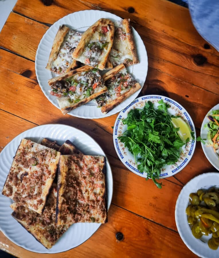 A wooden table in Avanos, Cappadocia, Turkiye, with various dishes including plates of cut flatbreads topped with meats and vegetables, a bowl of fresh herbs, and some