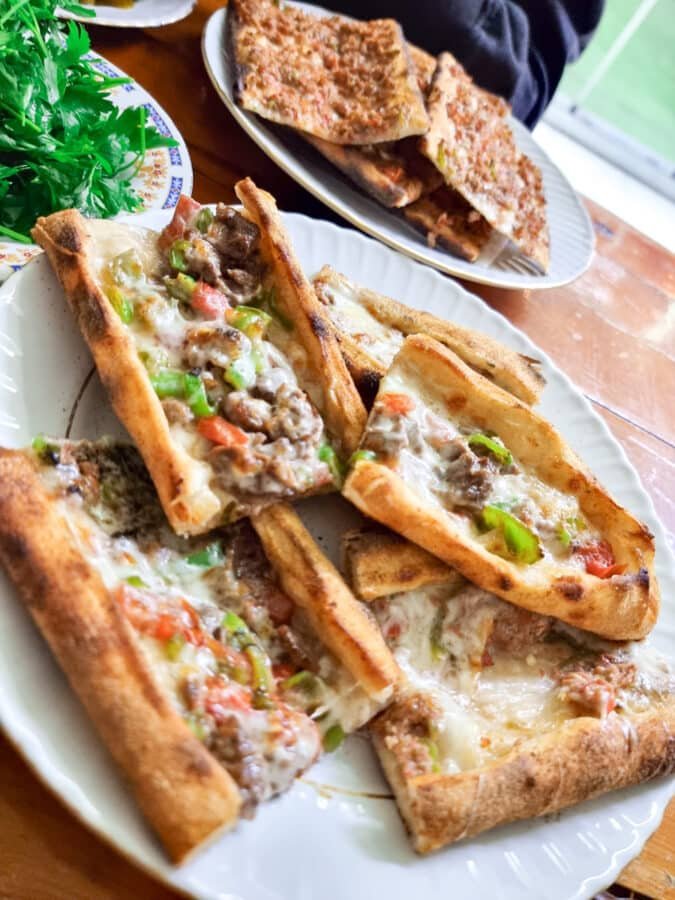 A close-up of a tray with slices of cheesy beef and vegetable pide, with a plate of garlic bread in the background, showcasing local cuisine from Avanos Cappadocia, Turkey.