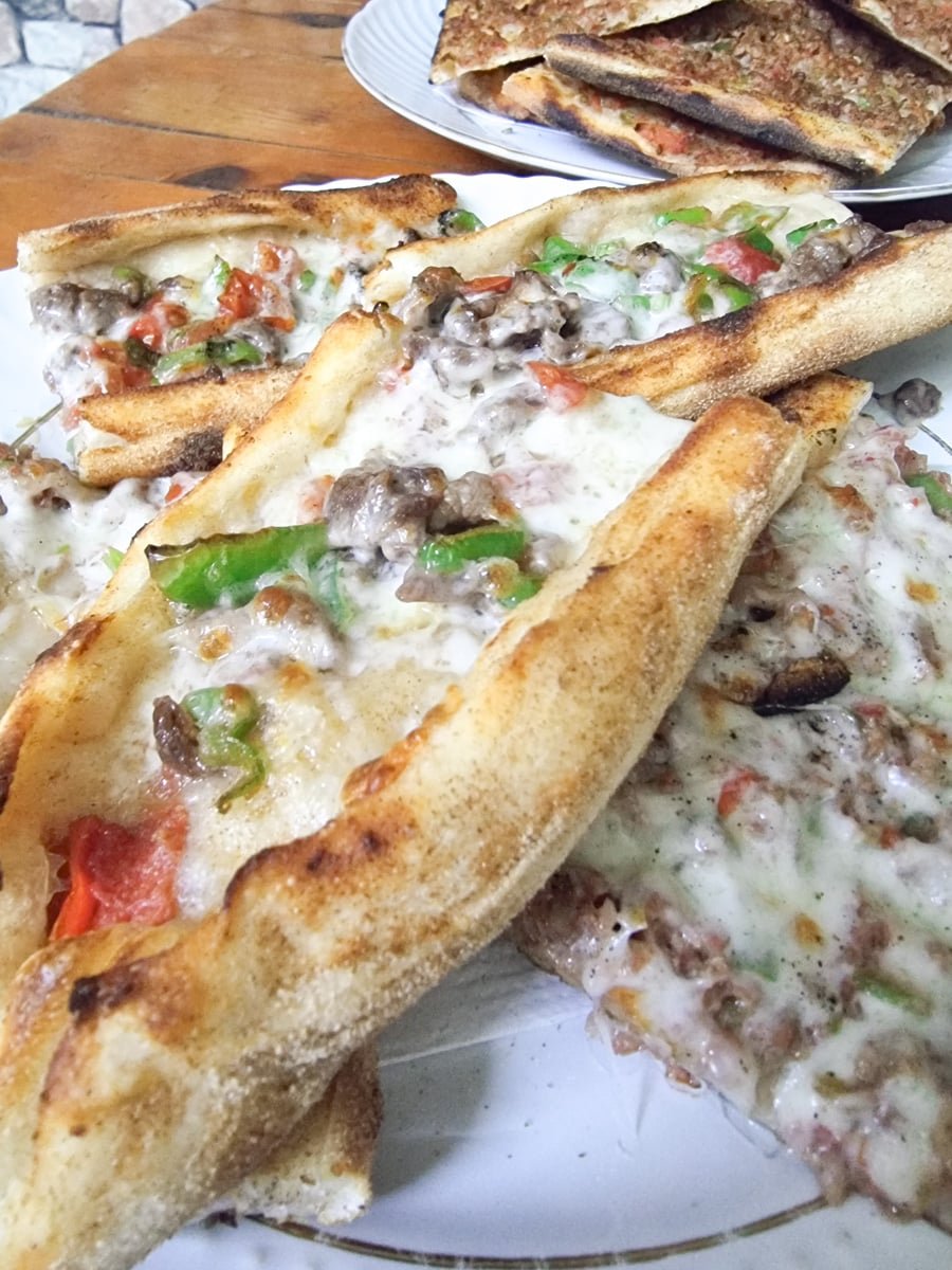 Plate of cheesy pide topped with vegetables and meat, served on a rustic wooden table in Avanos, Cappadocia, Turkey.
