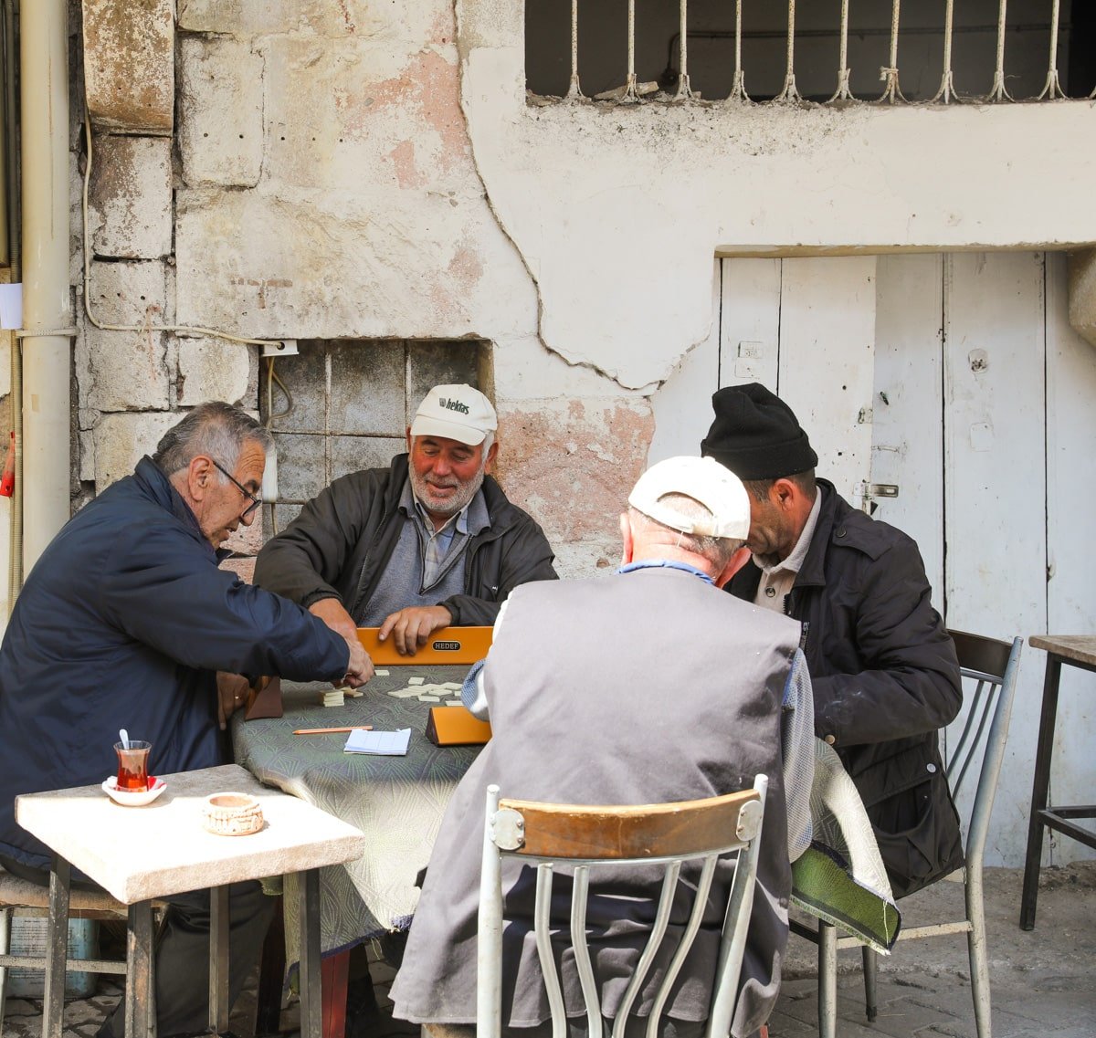 Four elderly men playing the board game Okey at an outdoor table near an old building wall in Avanos, Cappadocia, Türkiye, with one smiling broadly. Two are wearing caps.