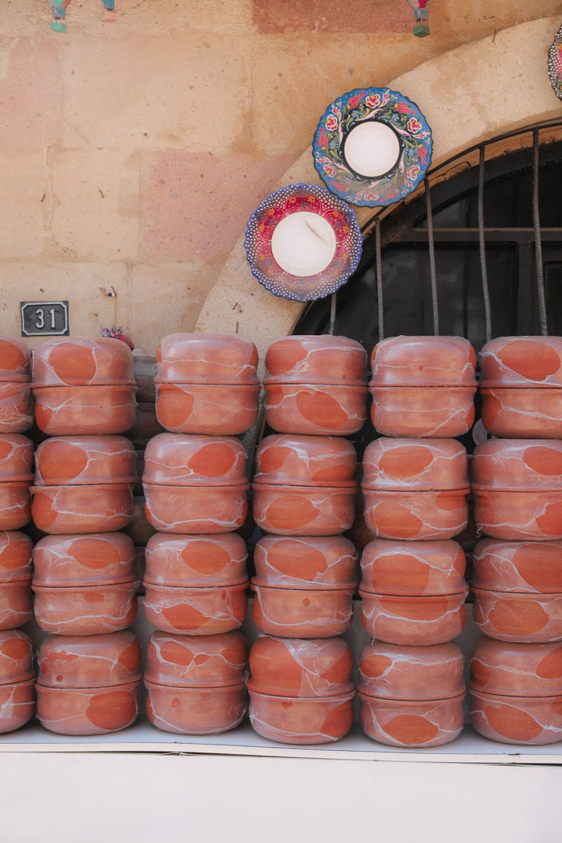 Stacks of traditional clay pots on a shelf outside a shop in Avanos, Cappadocia, Turkey, with colorful mandala plates hung on the wall above.