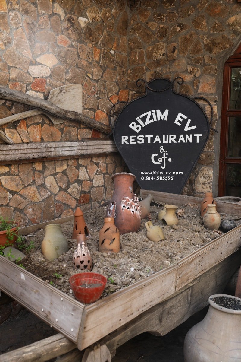 Sign for "bizim ev restaurant & cafe" mounted on a stone wall in Avanos, surrounded by an array of traditional clay pots and decorative items.