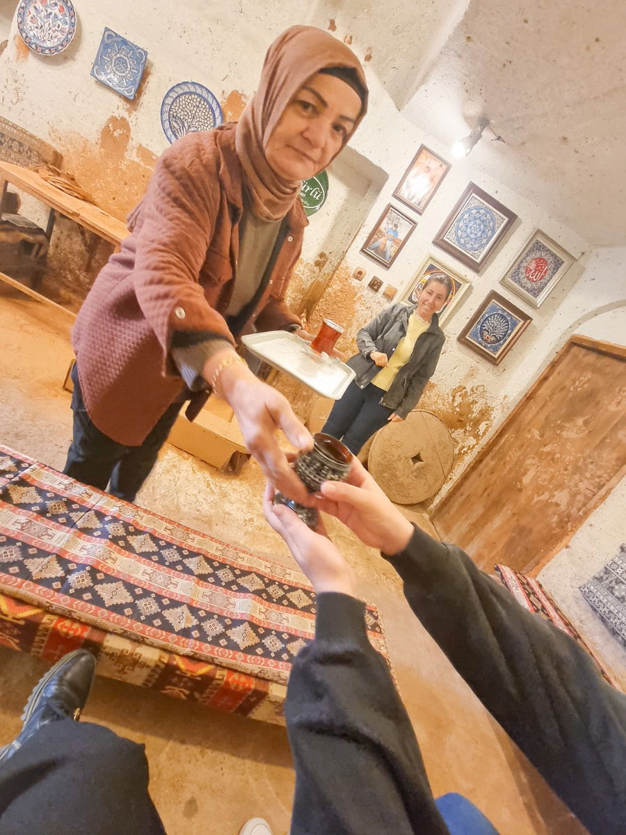 A woman with a headscarf handing a cup to a person in Avanos, Cappadocia, with another woman observing in a room decorated with ceramic plates.