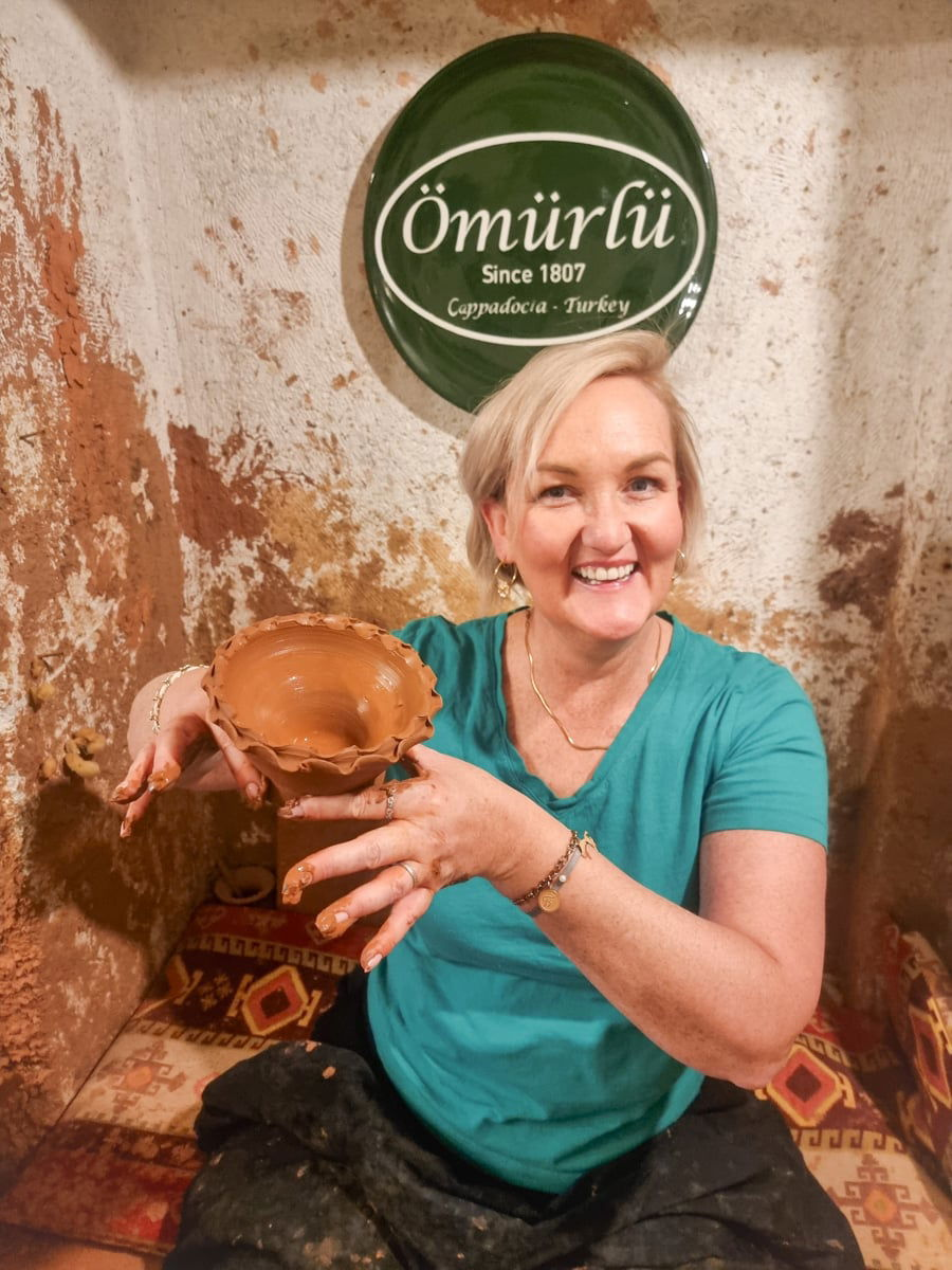 A smiling SJ in a teal shirt displays a freshly shaped clay pot in a pottery workshop, with a sign reading "Omirli" in the background, highlighting one of the things to do in Av