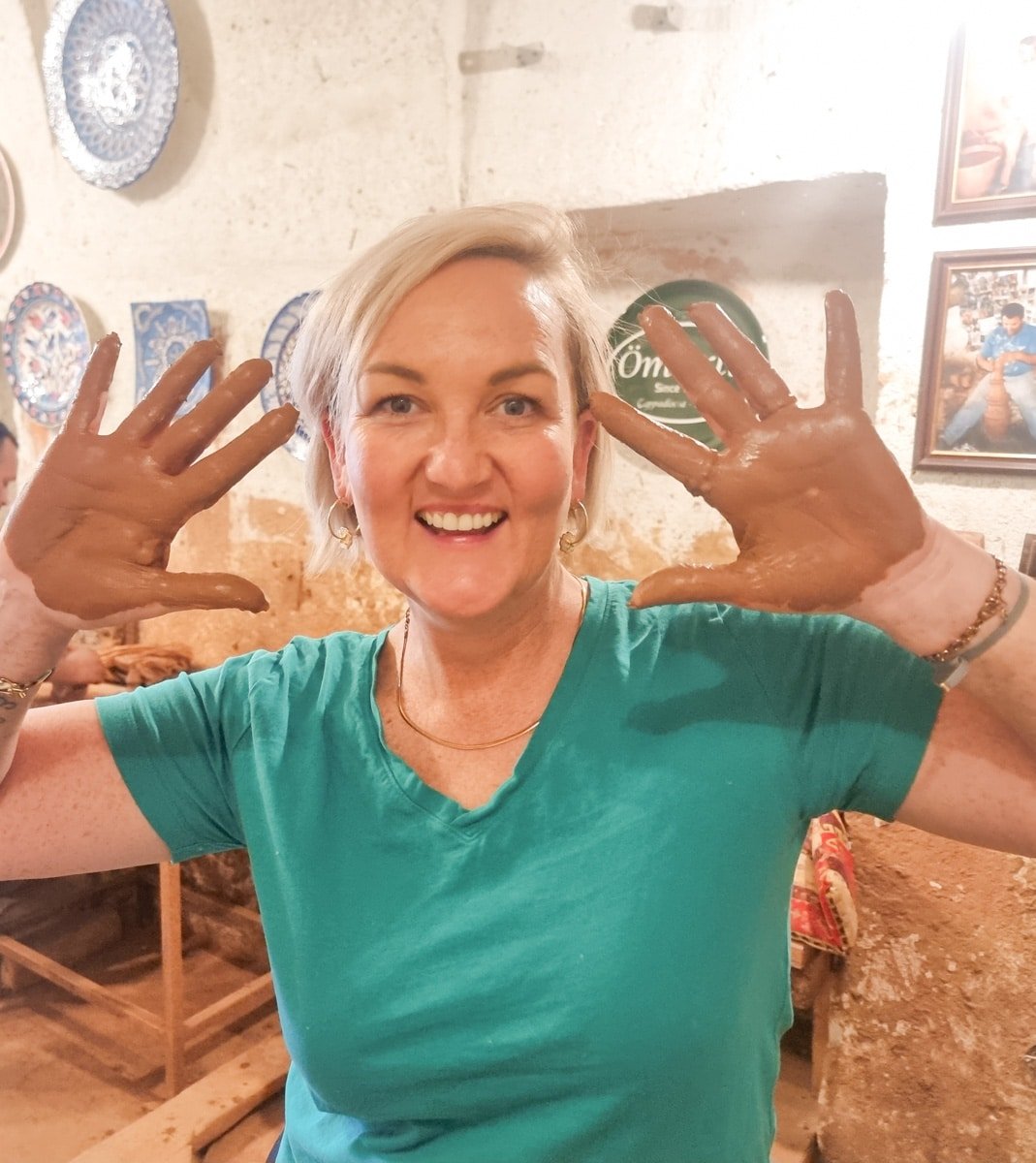 SJ with clay-covered hands smiles broadly inside a pottery workshop in Avanos, Cappadocia, Turkey, holding up her hands to display the clay.