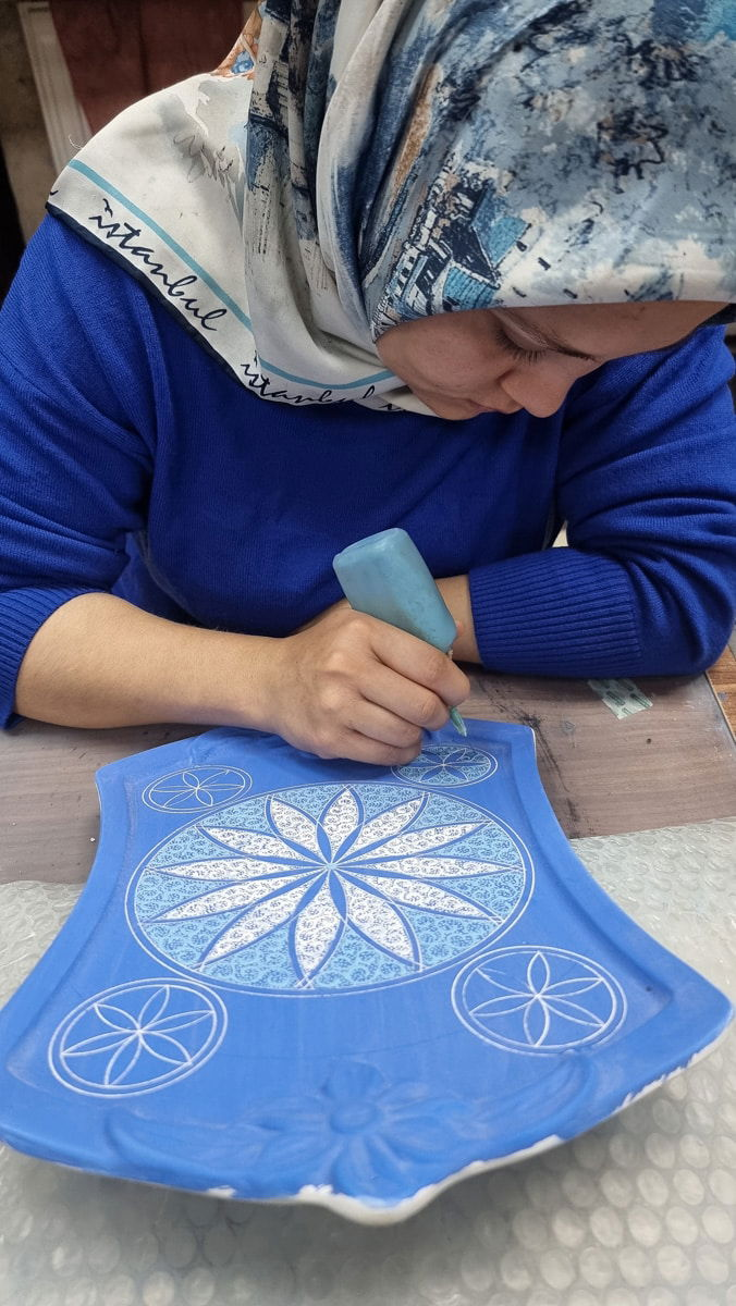 A woman in a patterned headscarf painting a blue cloth with a white floral design, focusing intently on her artwork in Avanos, Cappadocia, Turkey.