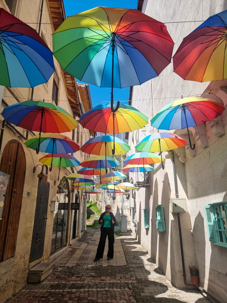 A person standing under a canopy of colorful hanging umbrellas in a narrow cobblestone street in Avanos, Cappadocia, Turkiye, with sunlight casting shadows on the walls.