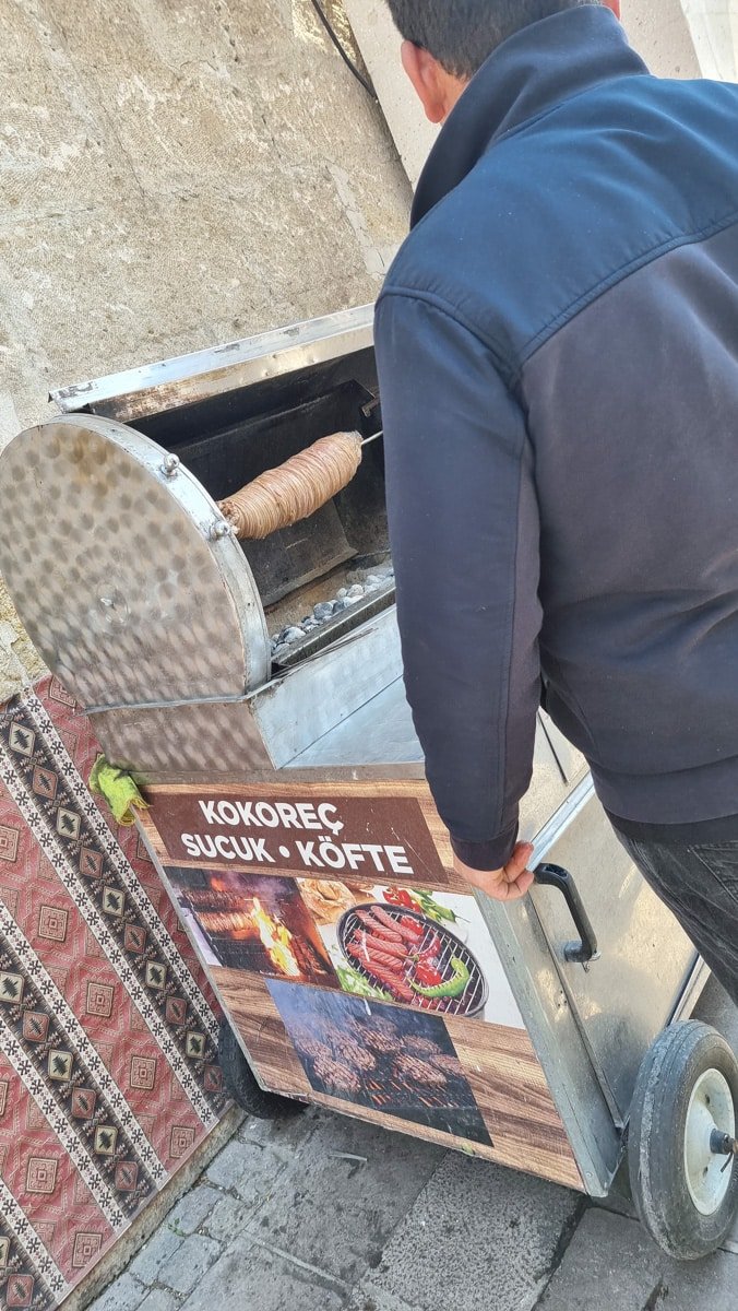 A man prepares traditional Turkish street food in a portable metal stove on a sidewalk in Avanos, Cappadocia, Turkiye, with a sign displaying "kokorec, sucuk,