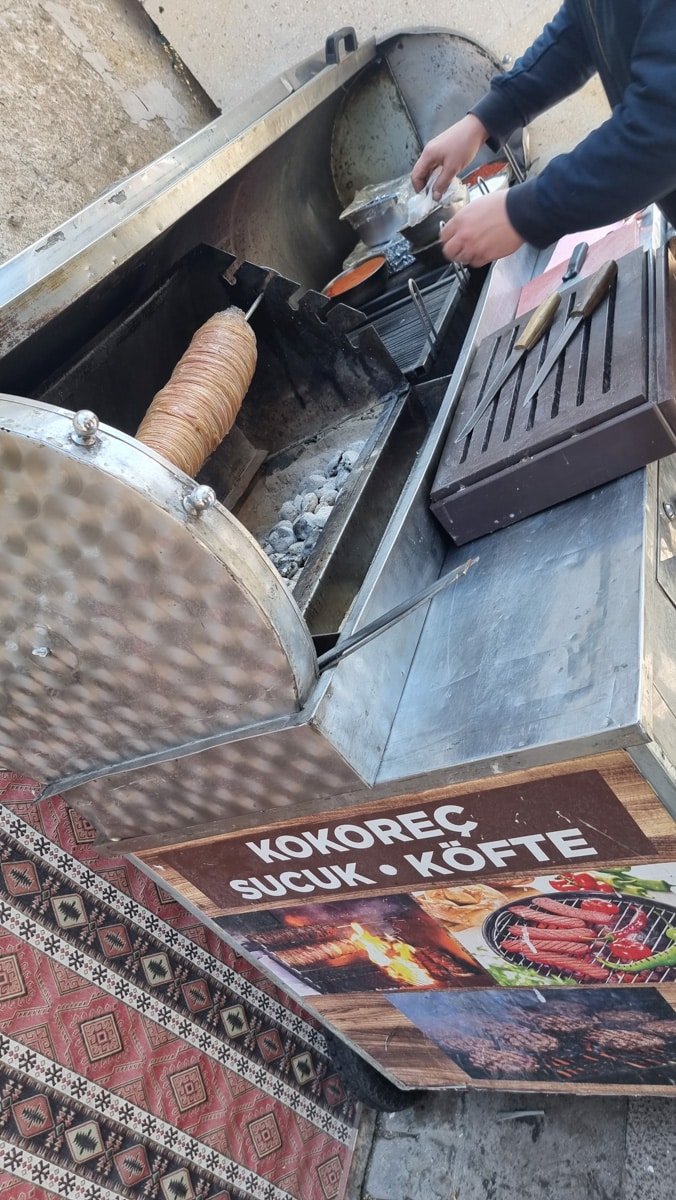 A vendor in Avanos Cappadocia, Turkiye, preparing street food, slicing warm kokoreç from a rotating spit over coals, with a sign displaying "kokoreç bü