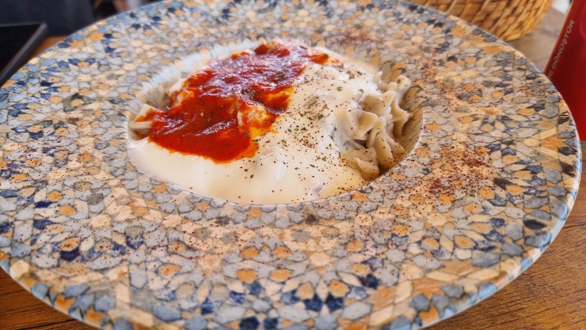 A plate of manti (Turkish dumplings) topped with garlic yogurt and spicy tomato sauce, garnished with dried mint, enjoyed in Avanos, Cappadocia, Türkiye.