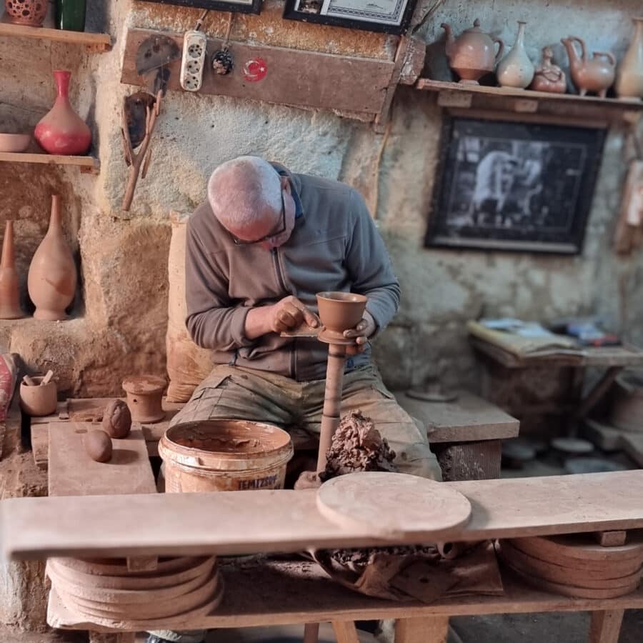 An elderly man crafting pottery on a traditional kick wheel in a rustic workshop surrounded by clay pots and tools, showcasing things to do in Avanos Cappadocia, Turkiye.