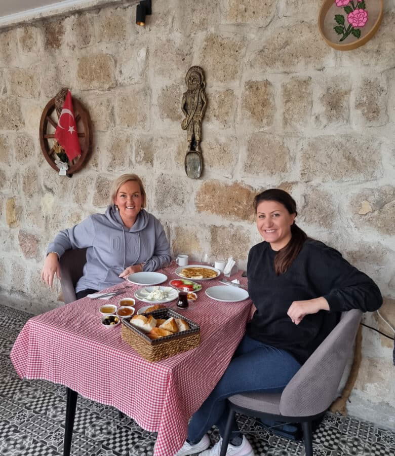 Two women smiling at a table with a traditional Turkish breakfast spread in a rustic stone-walled café in Avanos, Cappadocia.