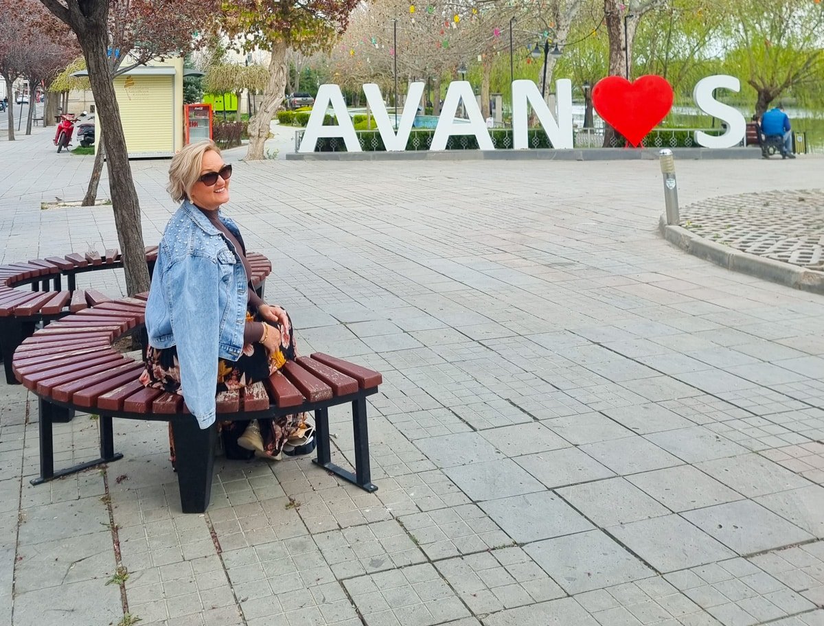 SJ IN sunglasses sitting on a circular bench in a plaza with a sign that reads "avans" featuring a red heart replacing the letter 'a', showcasing things to do in Avanos, Cappadocia