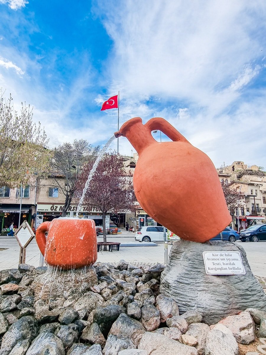 An outdoor fountain sculpture shaped like a large terracotta jug, expelling water, with a Turkish flag in the background and a plaque on a stone base, highlighting things to do in Avanos C