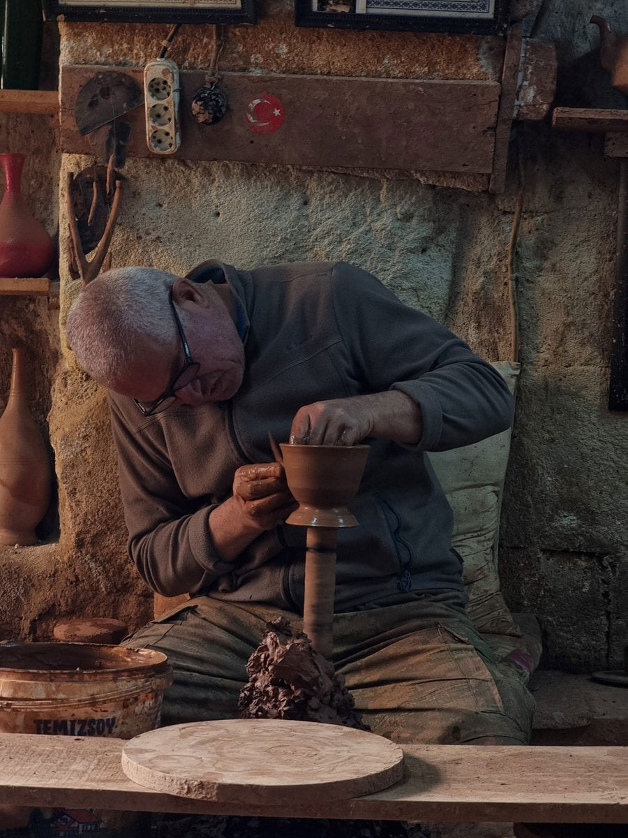 An elderly potter shapes clay on a pottery wheel in a rustic workshop in Avanos, Cappadocia, surrounded by tools and pottery pieces.