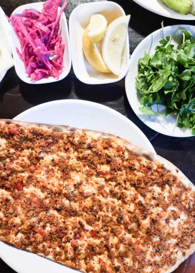 Rectangular Turkish lahmacun surrounded by sides of pickled cabbage, lemon slices, and fresh greens on a table at Ushiscar Castle in Cappadocia.