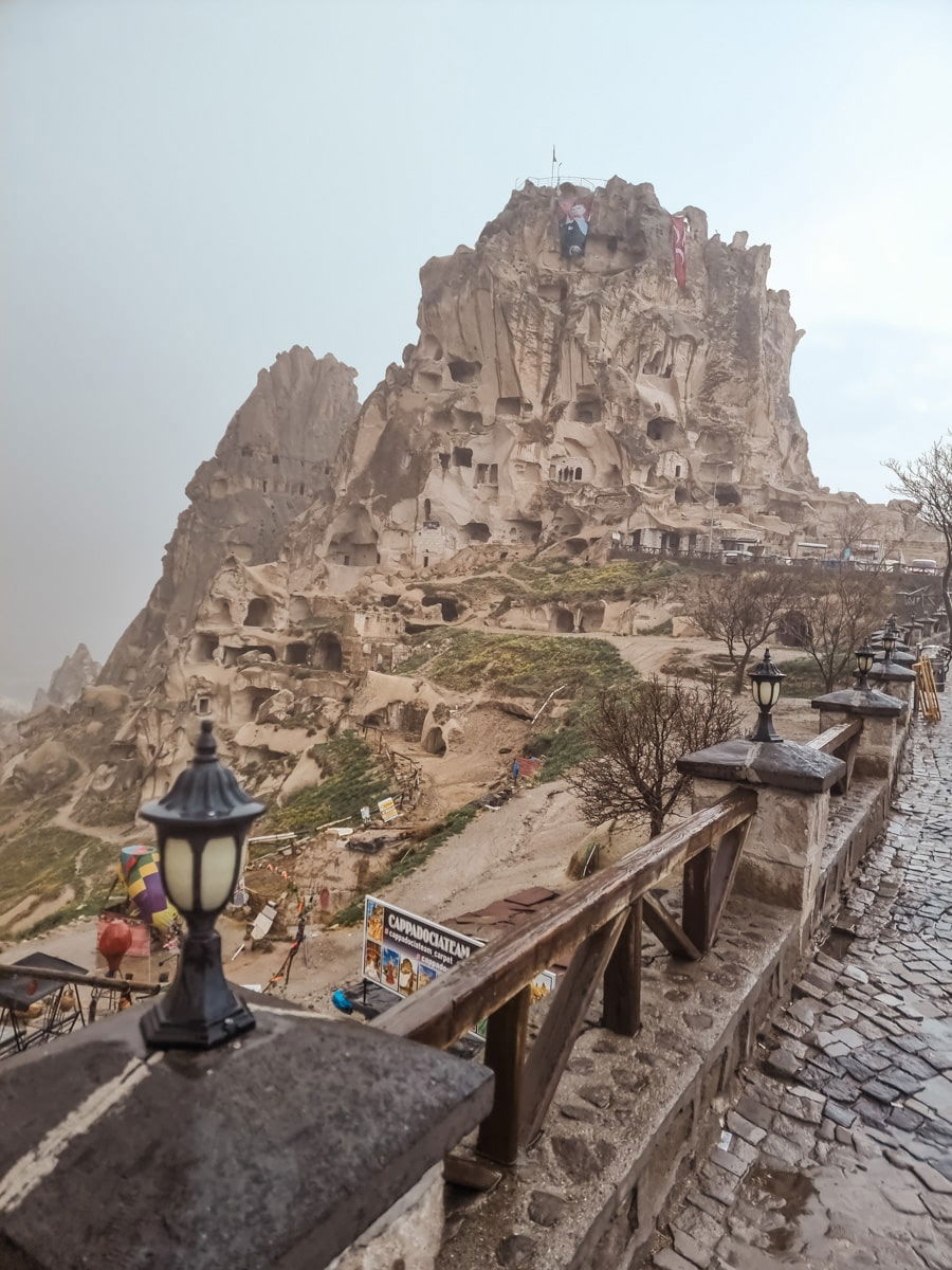 View of a foggy hillside with Uchisar Castle carved into a rocky landscape in Cappadocia, featuring a pathway lined with lamps. Uchisar on a rainy day