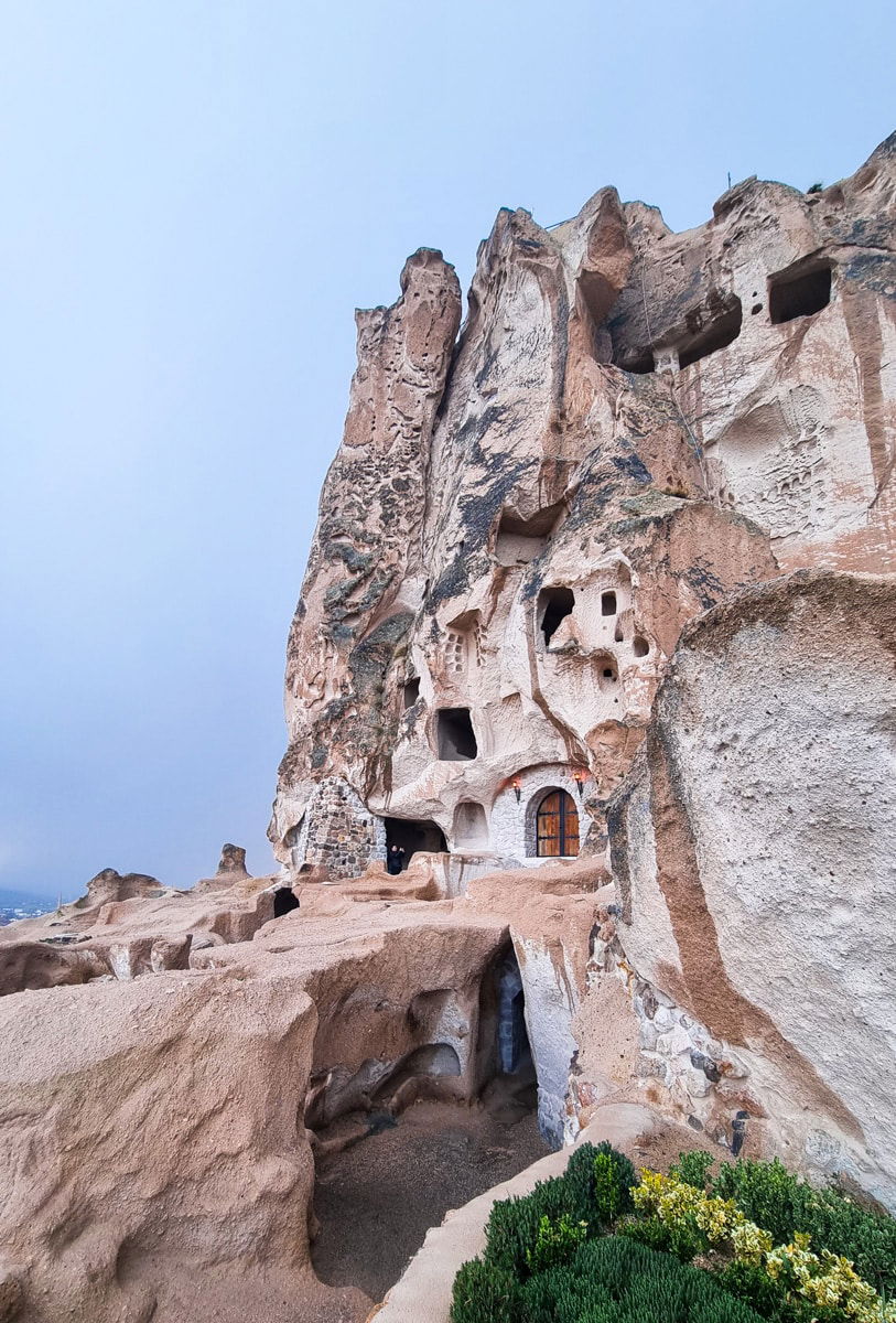 Ancient cave dwellings carved into a rocky hillside near Uchisar Castle in Cappadocia, Turkey, under a cloudy sky.