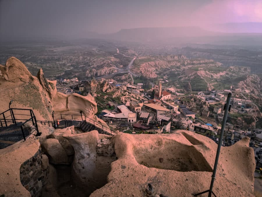 Aerial view of a village near Uchisar Castle in Cappadocia, Turkey, at dusk, featuring rock formations and rugged terrain with a river in the background.