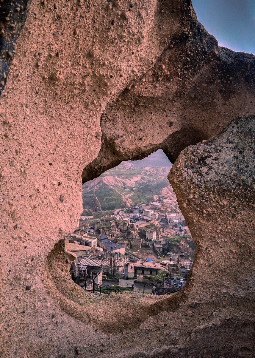 A scenic view through a heart-shaped natural opening in a rock formation, overlooking the quaint Ushiscar Castle nestled among lush hills at sunset.