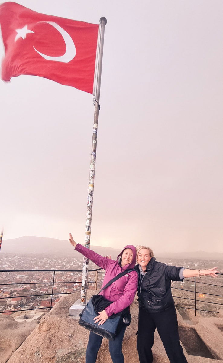 Two women smiling and posing with outstretched arms under a large Turkish flag on a hilltop near Ushisar Castle in Cappadocia at dusk.