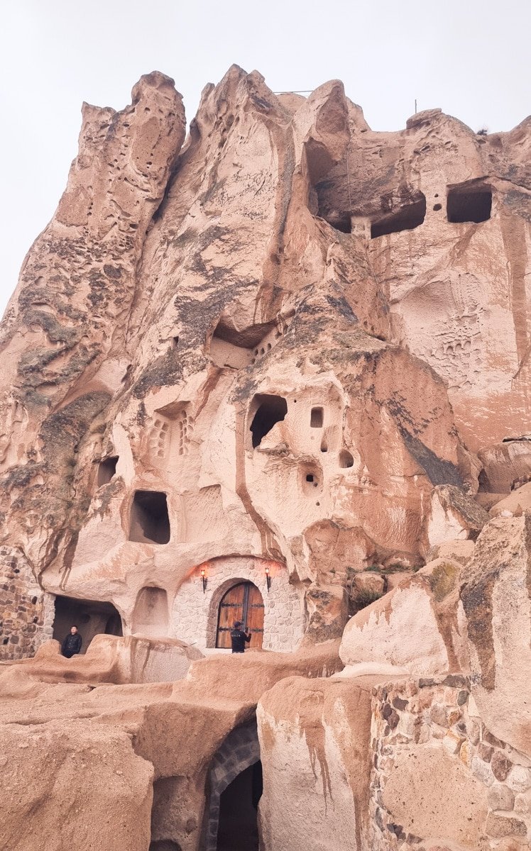 Ancient cave dwellings carved into a beige rock formation with multiple openings and a visible door, located near Uchisar Castle in Cappadocia, Turkey.