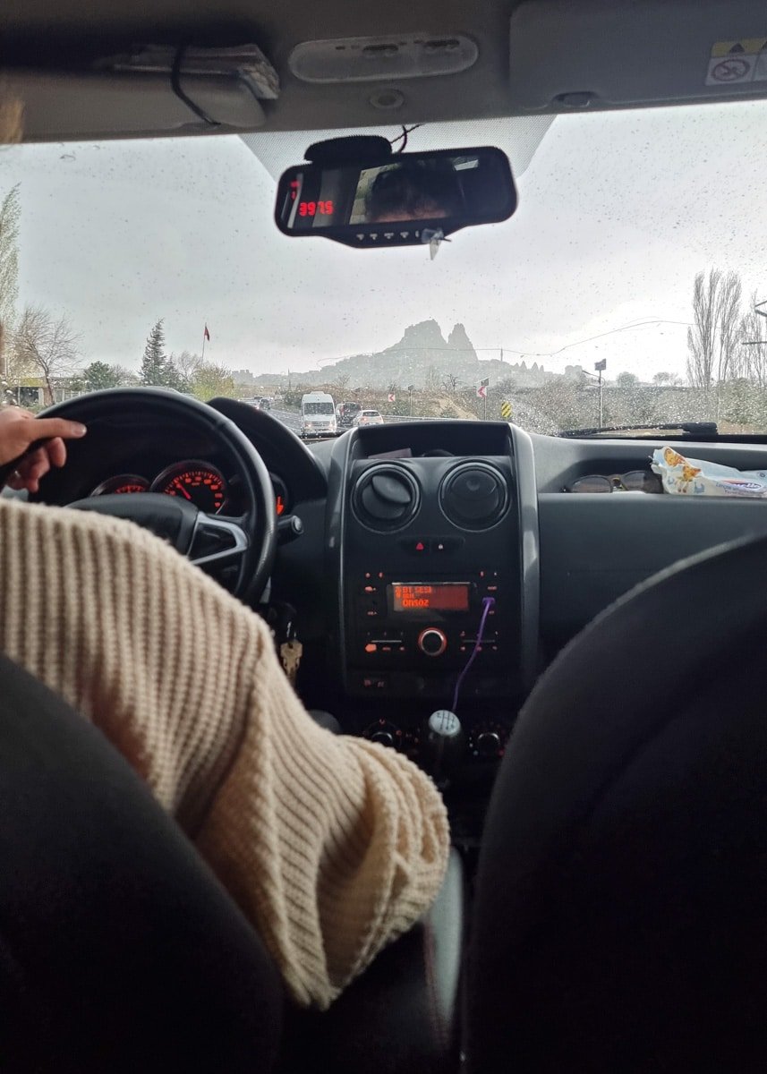 View from the backseat of a car showing the dashboard and steering wheel, with a road leading towards the distant Ushiscar Castle in Cappadocia under a cloudy sky.