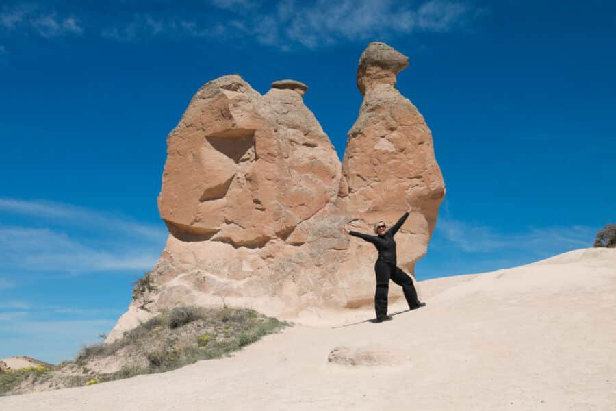 A person posing with their arms raised in front of two large, uniquely shaped rock formations in Devrent Imagination Valley, Cappadocia, under a clear blue sky.