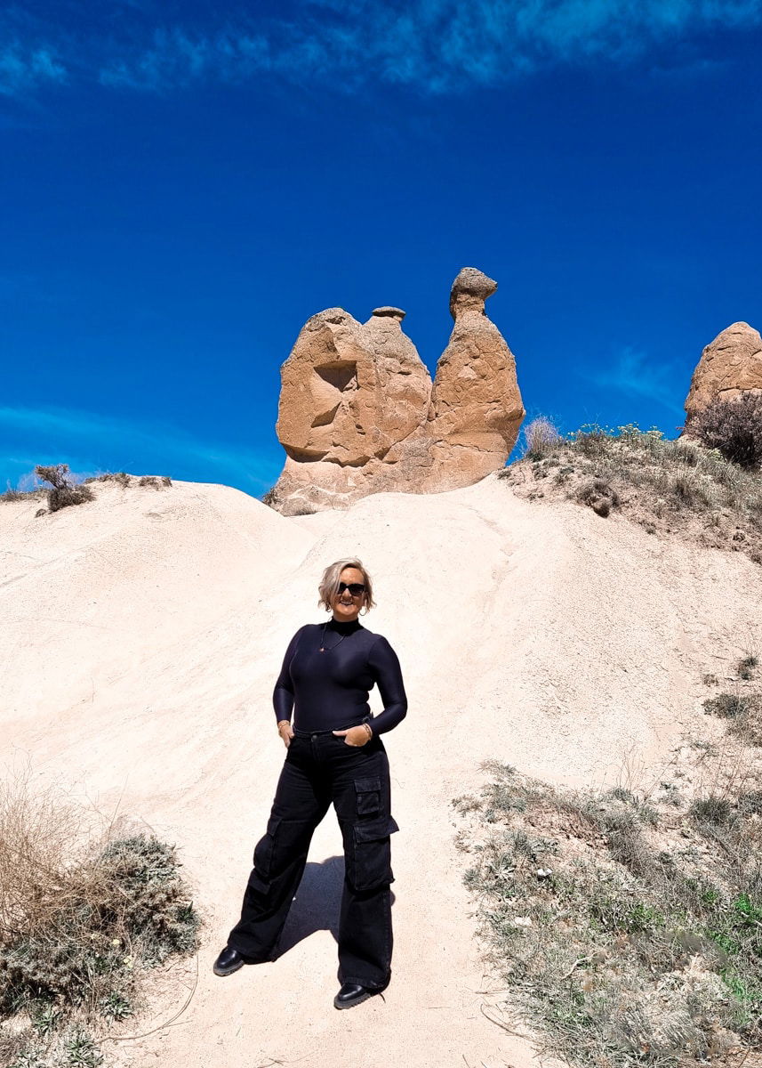 SJ standing on a sandy trail in Devrent Imagination Valley, Cappadocia, with unique rock formations in the background under a clear blue sky.