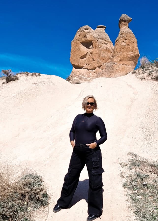 A woman in a black outfit stands in front of a large, uniquely shaped rock formation in Devrent Imagination Valley, Cappadocia, under a clear blue sky.