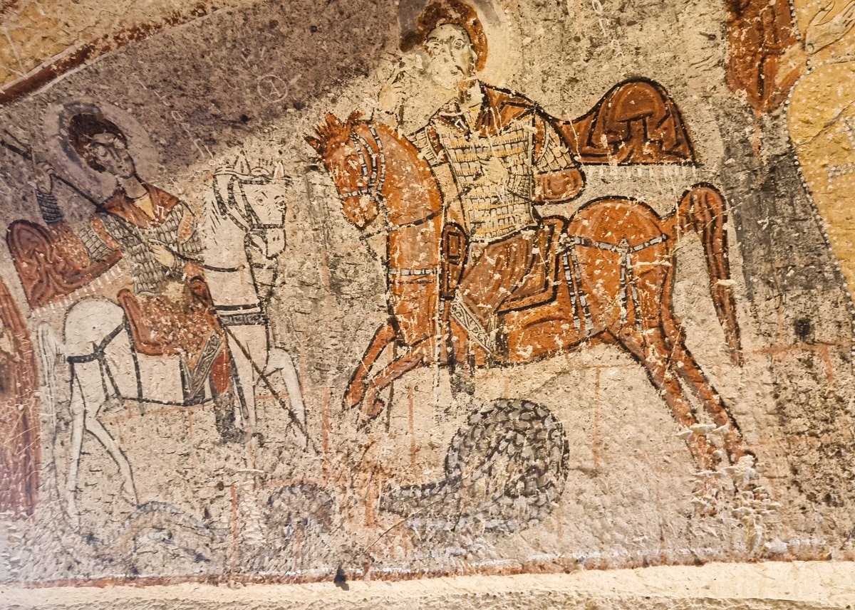 Ancient fresco depicting a warrior on a horse with an accompanying musician playing a lyre, painted in earthy tones on a rough textured wall at the Goreme Open Air Museum.