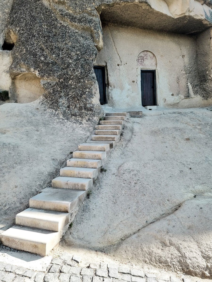 Stone steps leading up to a door carved into a rocky hillside at the Goreme Open Air Museum with another wooden door to the left.