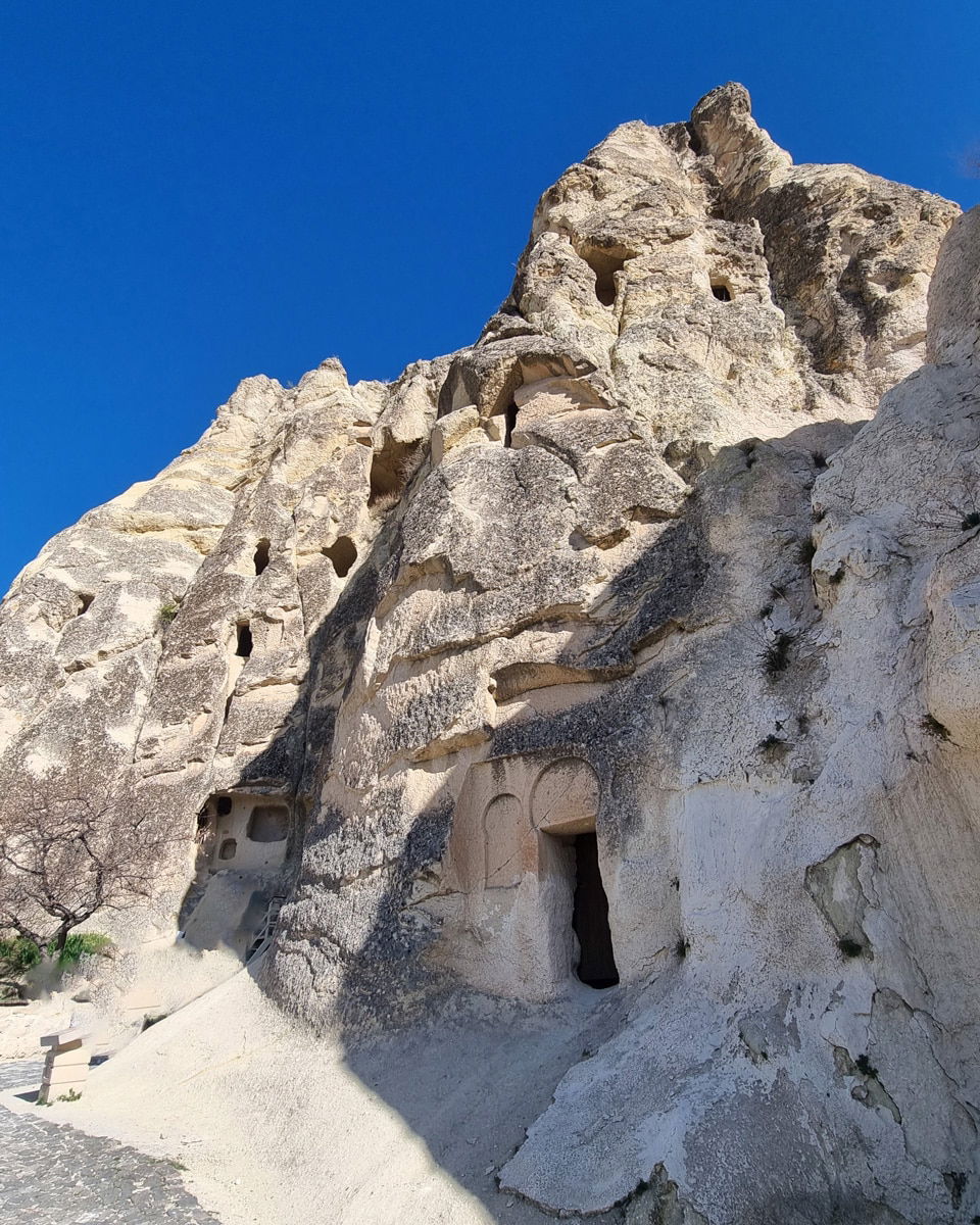 Rock formations and ancient cave dwellings carved into a rocky hillside in the Goreme Open Air Museum, under a clear blue sky.