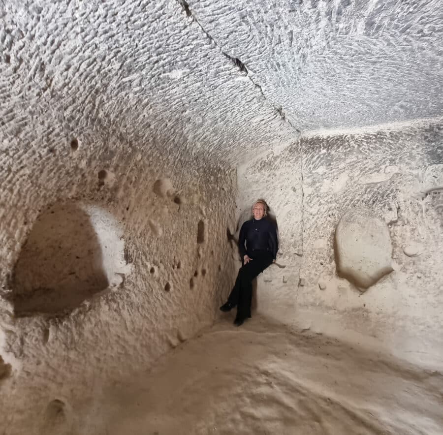 A person stands inside a spacious, carved area of the Ozkonak underground city with textured walls and dim lighting.