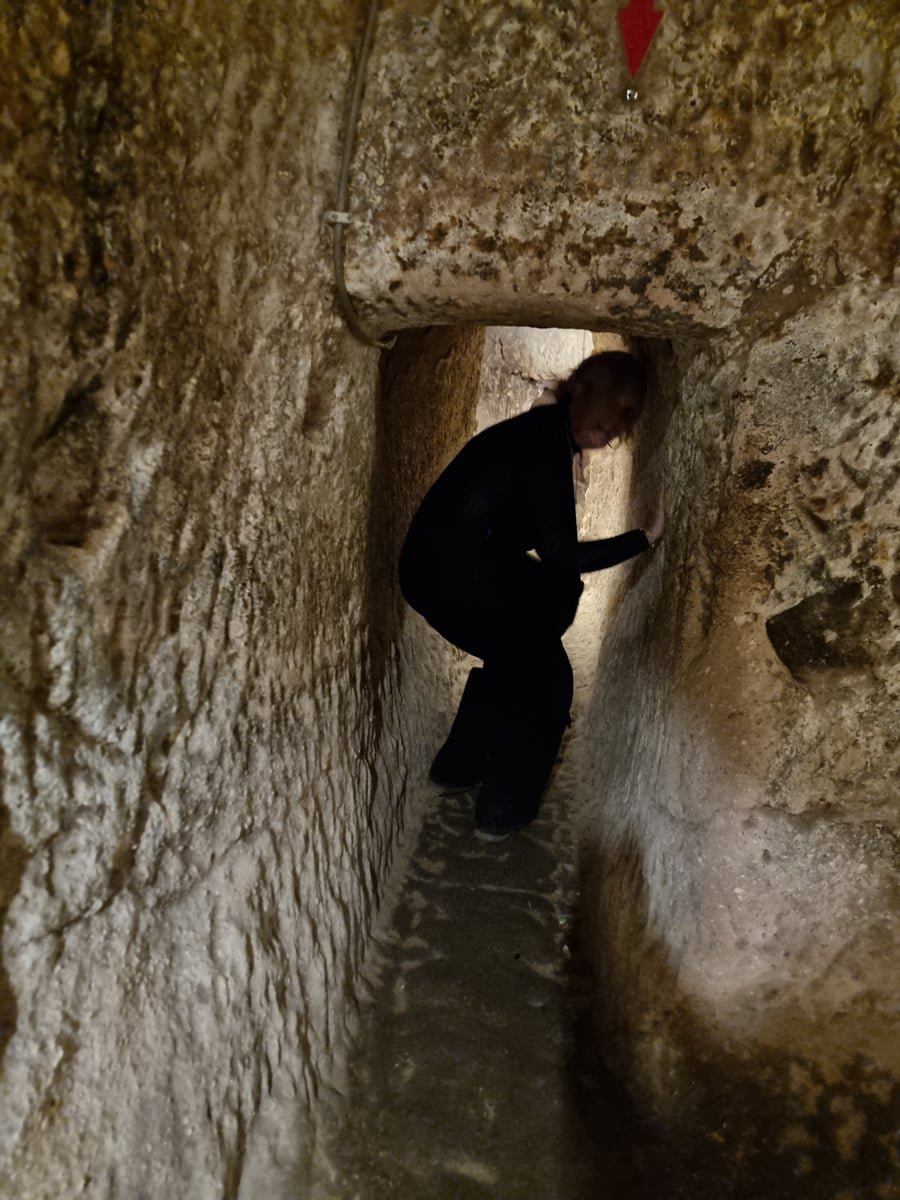 SJ bending slightly while walking through a narrow, dimly lit stone corridor in Ozkonak Underground City, with an arrow sign pointing downward.
