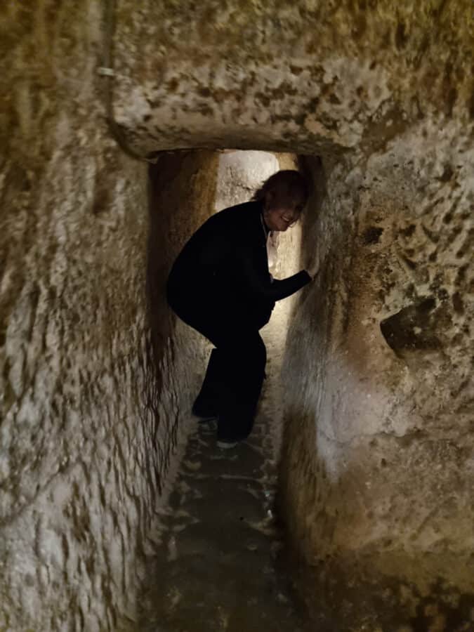 A person in black clothing navigates through a narrow, dimly lit tunnel in Ozkonak Underground City, crouching slightly to fit in the cramped space.