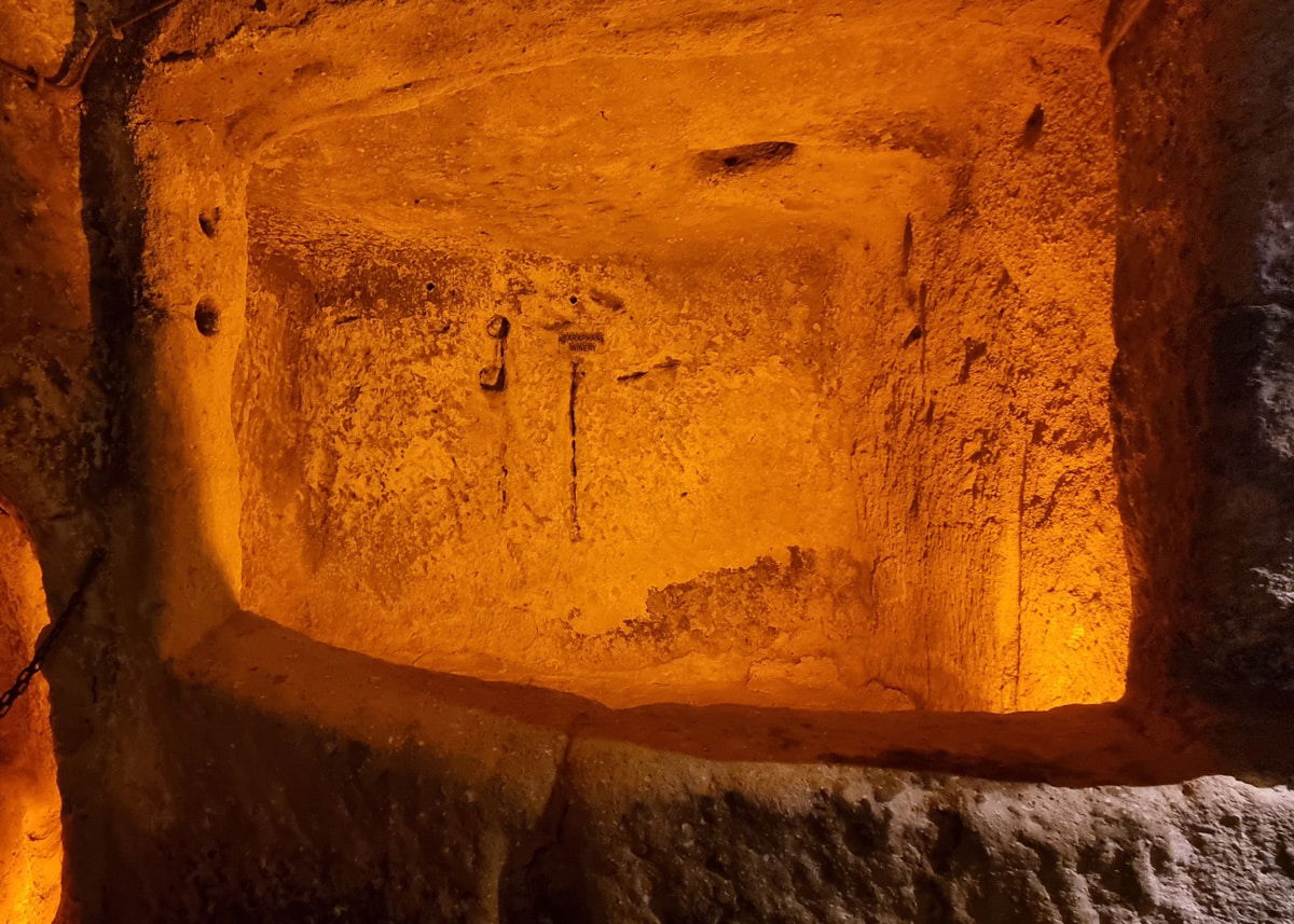 A warmly lit underground cave within Ozkonak Underground City, with textured stone walls and floor, illuminated by orange light creating deep shadows.
