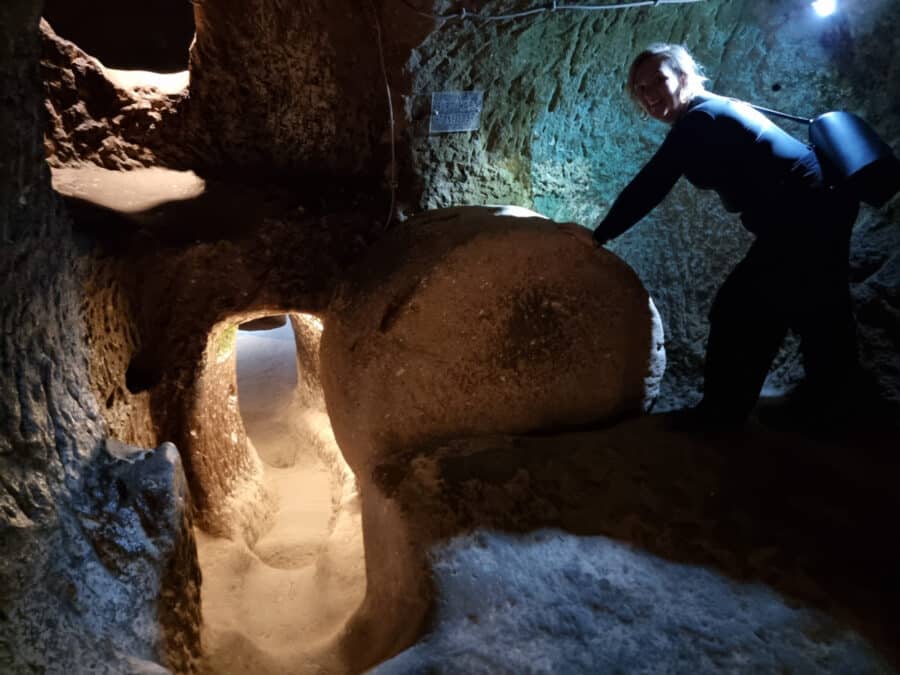 A person exploring a dimly lit tunnel in Ozkonak underground city, smiling at the camera, standing beside a large carved stone.