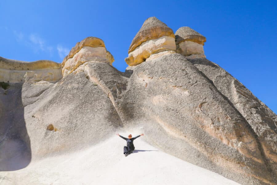 Person with arms raised standing on white rocky hill in Pasabag Valley, Cappadocia, Turkey, with tall, sculpted rock formations under a clear blue sky.