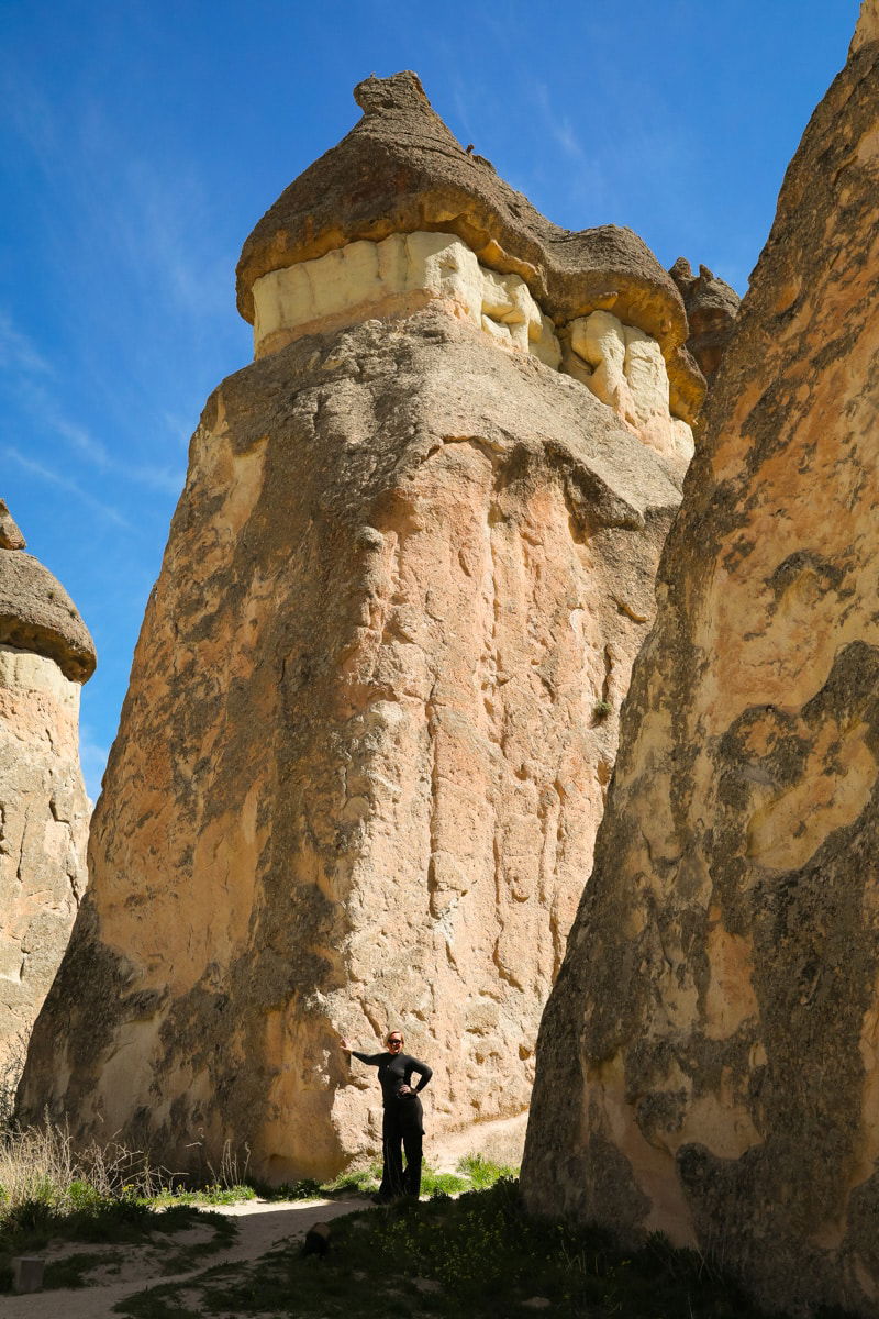 A person with arms raised stands beneath towering, uniquely shaped rock formations in Pasabag Valley, Cappadocia, under a clear blue sky.