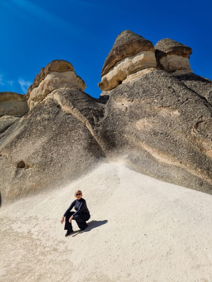 SJ sitting at the base of tall, uniquely shaped rock formations in Pasabag Valley, Cappadocia, under a clear blue sky.