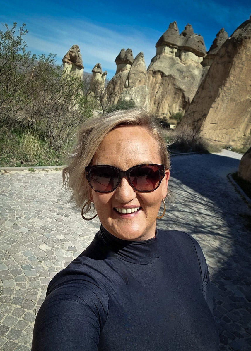 A woman in sunglasses takes a selfie with unique rock formations in Pasabag Valley, Cappadocia, in the background on a sunny day.