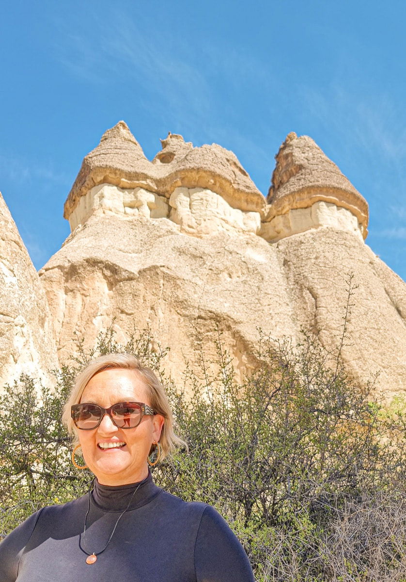 SJ with sunglasses stands in front of unique, conical rock formations in Pasabag Valley, Cappadocia, under a clear blue sky.