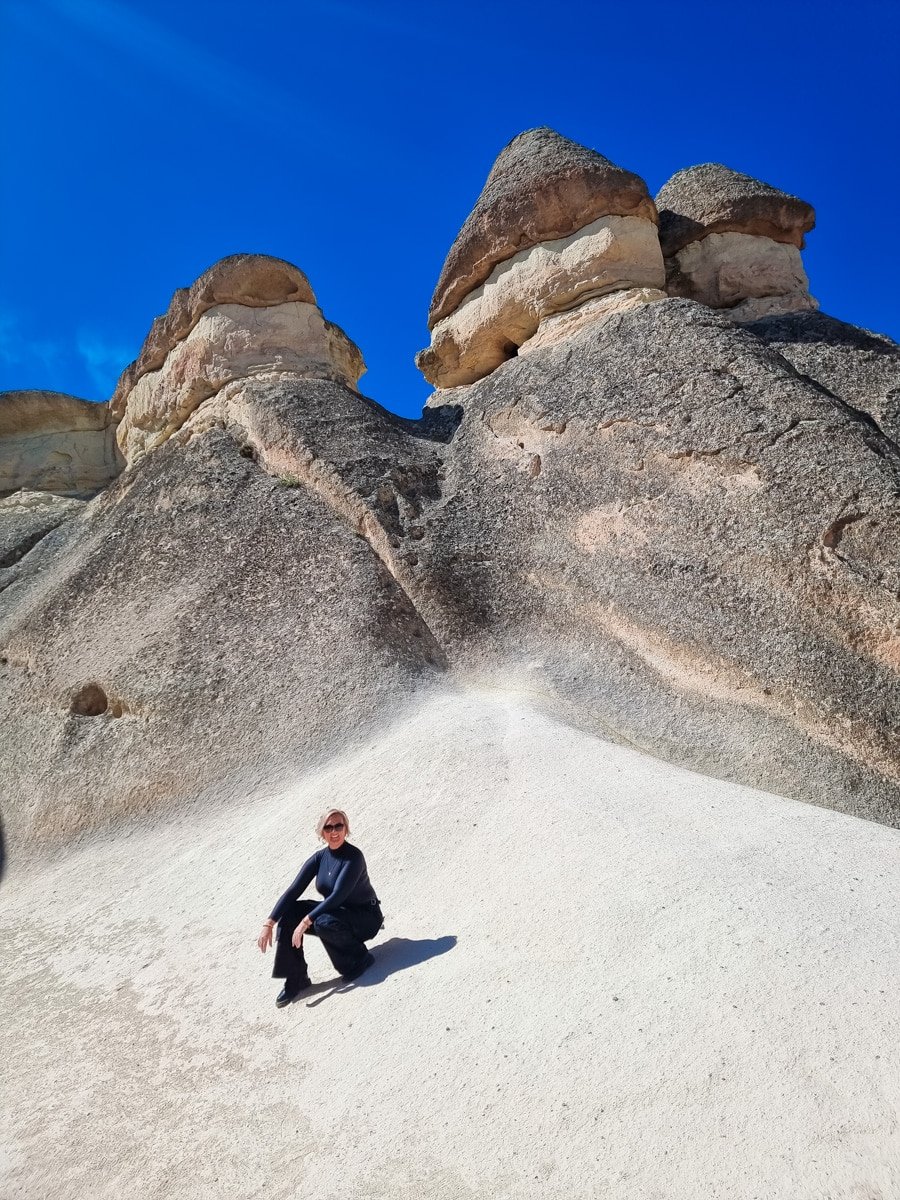 SJ squatting at the base of towering, uniquely shaped rock formations in Pasabag Valley, Cappadocia, under a clear blue sky.