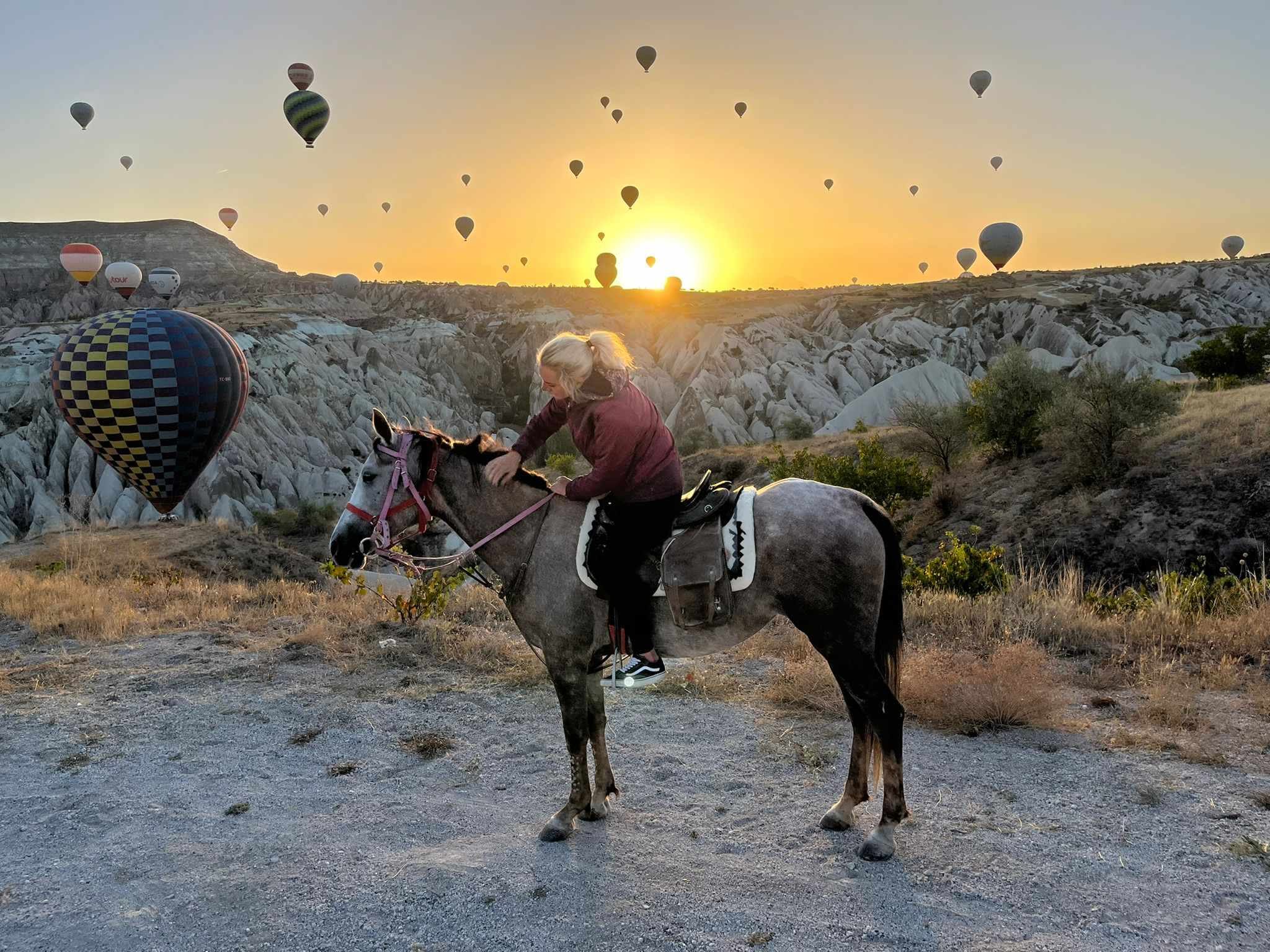 A person on a horse with another horse beside them at sunrise, with multiple hot air balloons in the sky above a rocky landscape.