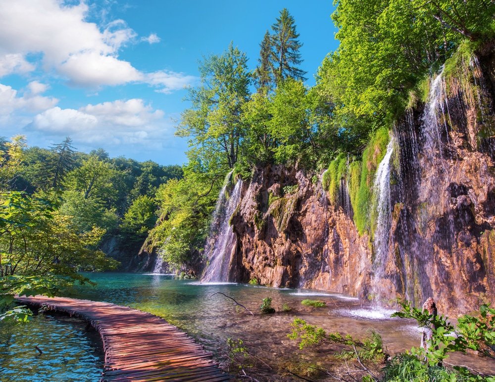 Scenic view of sunlit waterfalls cascading into a turquoise lake at Plitvice Lakes Entrance 1, with a wooden walkway surrounded by lush forest.