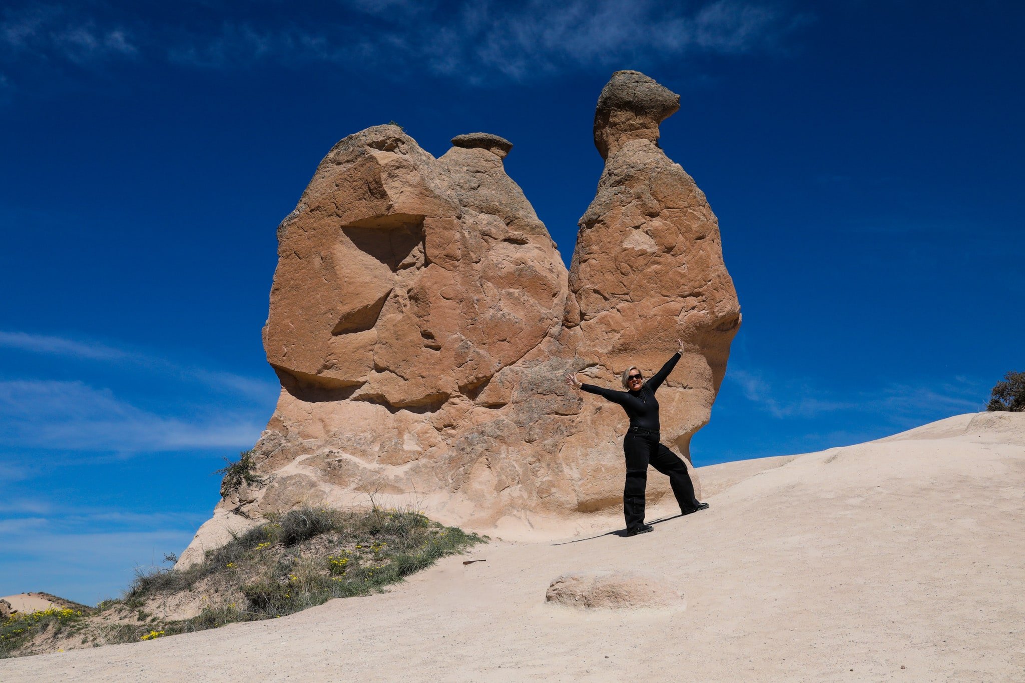 SJ posing with outstretched arms in front of unique, large rock formations under a clear blue sky during a Cappadocia Itinerary.