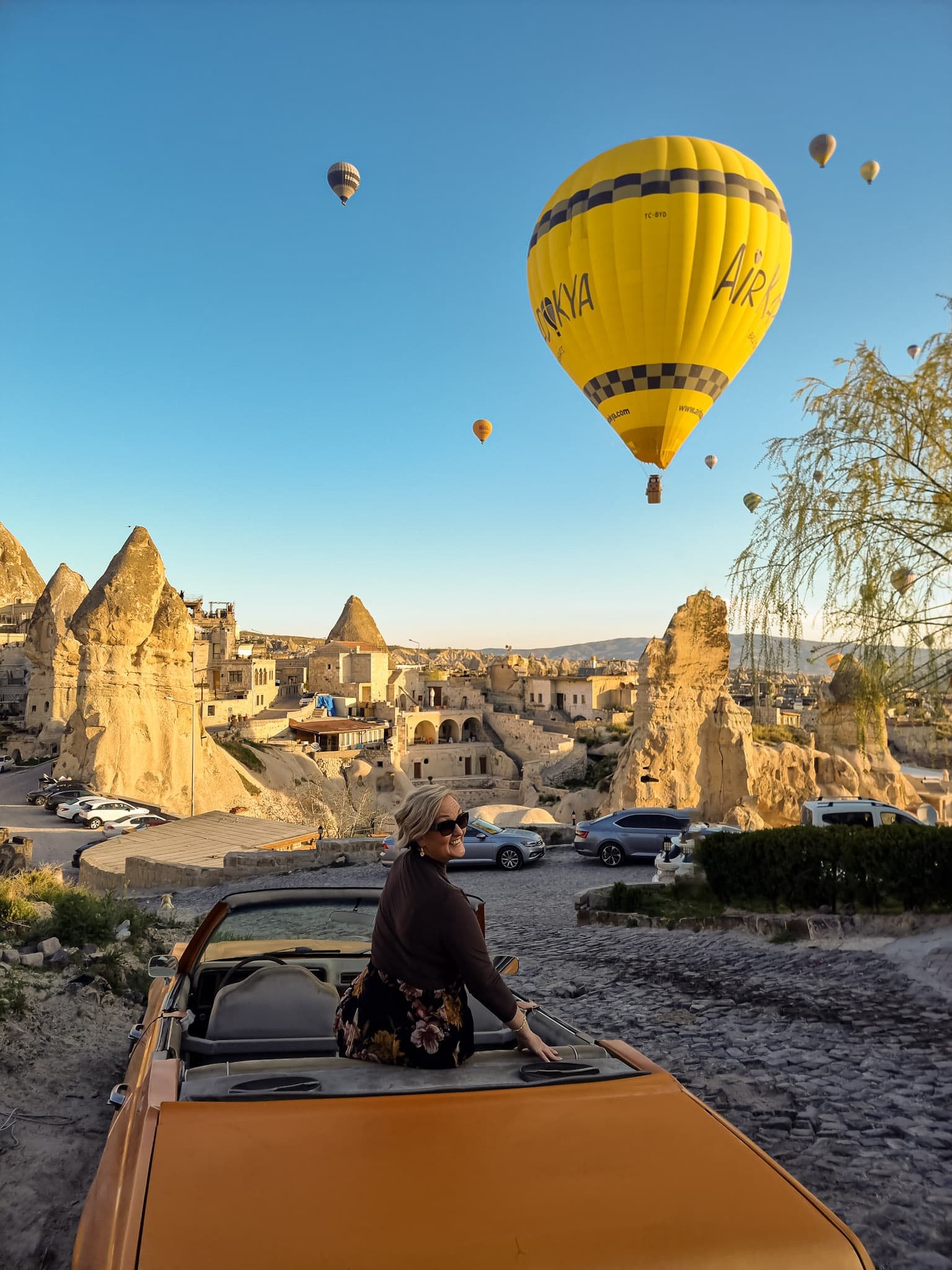 SJ sitting on a yellow vintage car watching hot air balloons fly over rocky landscape after 3 days in Cappadocia, Turkey.
